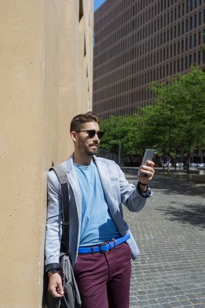 Bearded man in blazer leaning on a building wall outside with bag checking phone