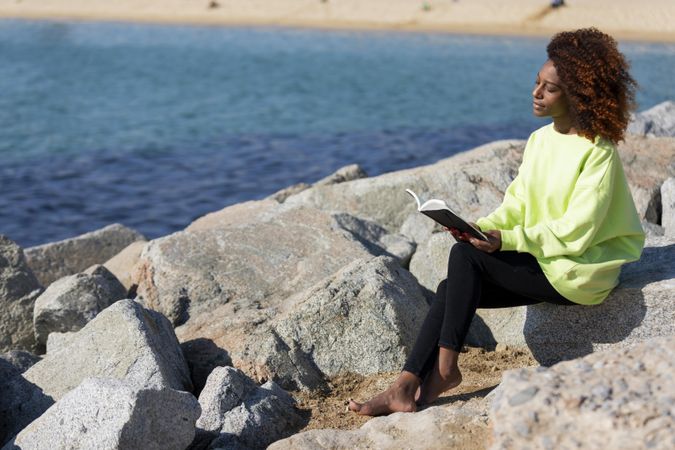 Female in bright green shirt sitting on the coast enjoying the sun with a book
