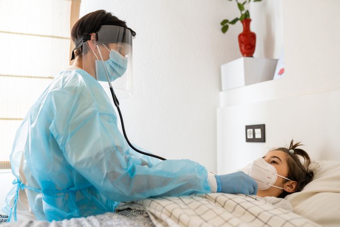 Doctor with facemask using stethoscope on a patient in bed