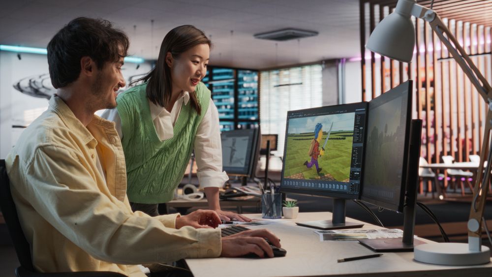 White Male Game Developer Using Desktop Computer With 3D modelling Software And Talking to Asian Female Project Manager in Game Design Studio Office.