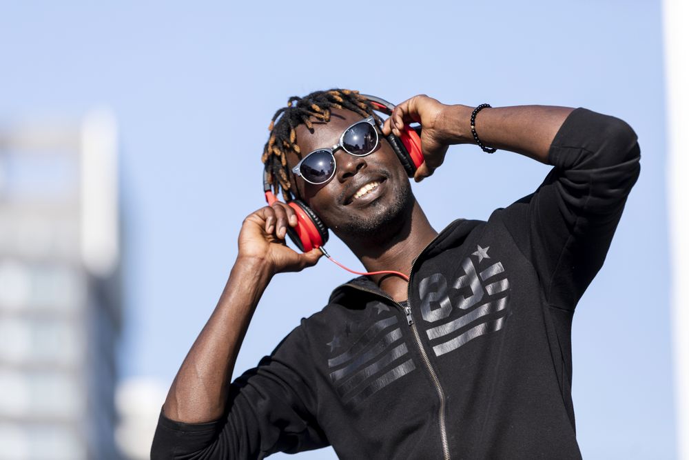 Smiling Black man wearing headphones & sunglasses standing on the street listening to music