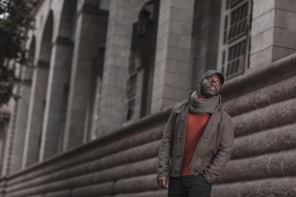 Man in brown jacket  and scarf standing beside a building