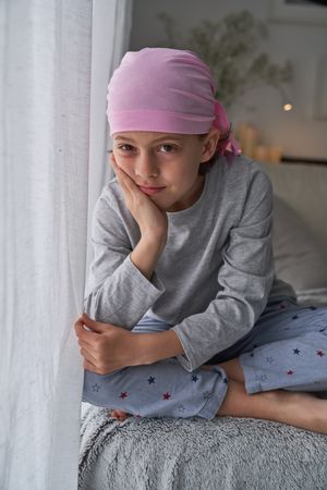 Cute boy in pink headscarf sitting near curtain