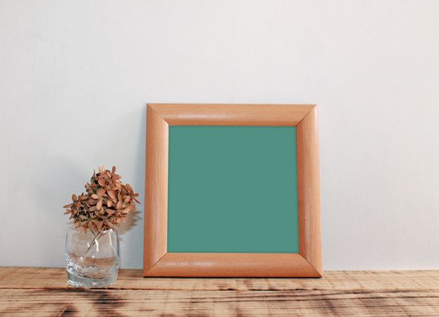 Plain square wooden picture frame with green interior leaning against wall mockup with cup of dried flowers