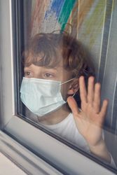 Cute boy in mask looking out of window at home