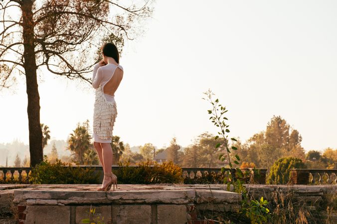 Back view full length shot of woman in formal white dress standing outside looking at view of trees