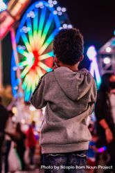 Young boy is grey jacket standing in front of colorful Ferris wheel 5lWWY0