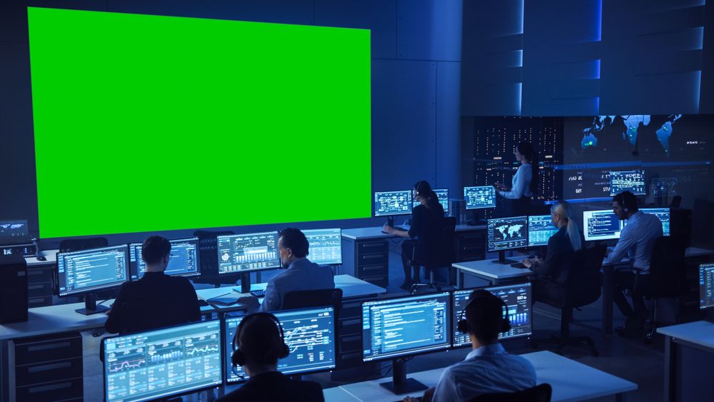 Diverse Team Monitors Data in Futuristic Command Center. Large Green Screen Dominates, Surrounded by Analysts at Workstations.
