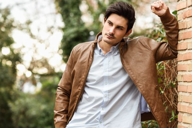 Mid shot of man in smart winter jacket and button up shirt leaning on brick wall outside