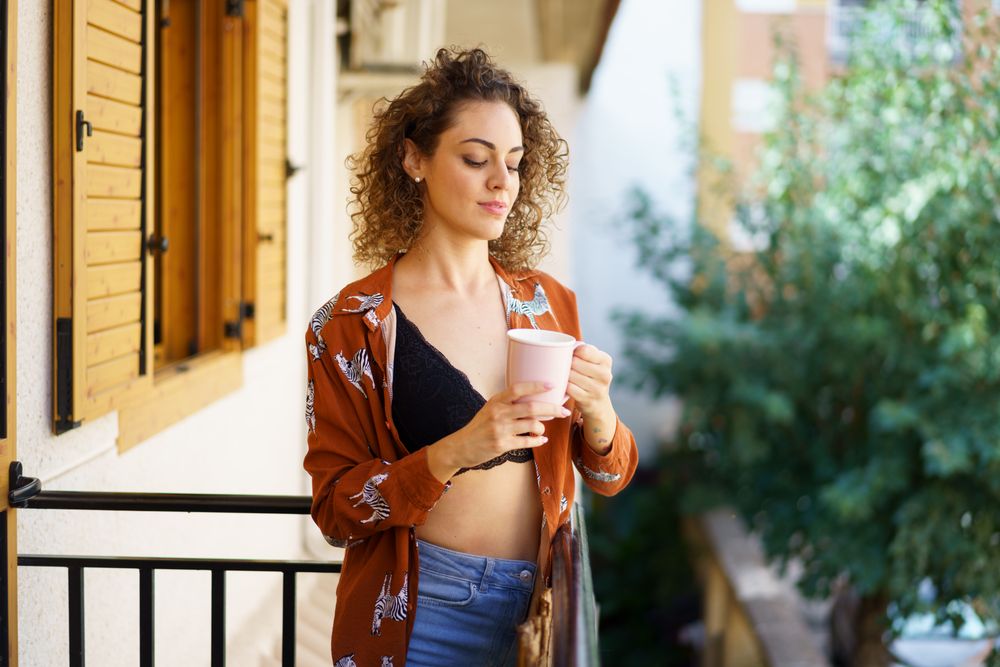 Young stylish smiling woman with cup of coffee standing on balcony at home