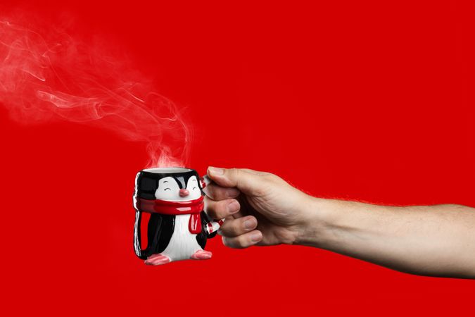 Hand holding a steaming penguin Christmas mug on a red background.