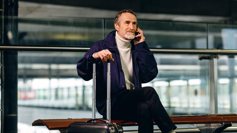 Older man sitting on bench outside of airport with suitcase using his cell phone