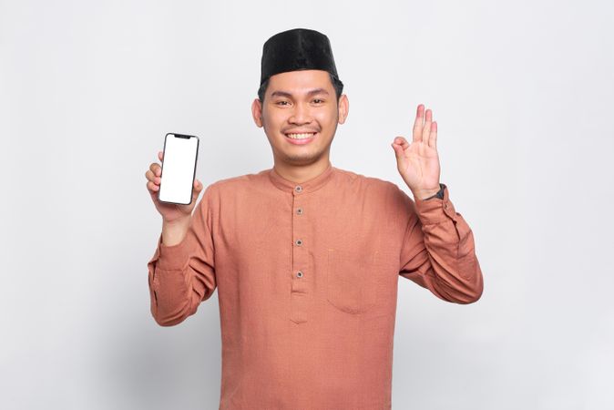 Muslim man in kufi hat smiling with mobile phone making a-ok sign with other hand