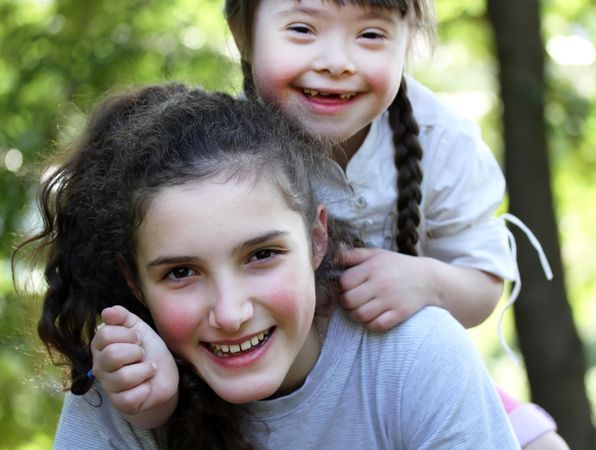 A young girl with Down syndrome on her teenage sister’s back in the park