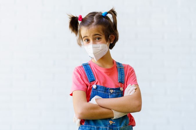 Cute child in overalls, gloves and facemask with arms crossed