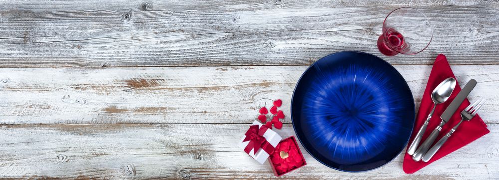 Table setting for Valentines Day celebration in flat lay format with blue plate, red hearts, napkin, silverware and drinking glass on rustic wood