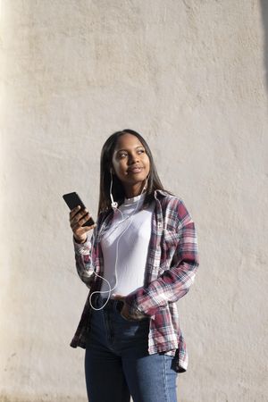 Female standing in the sun in front of wall holding phone and looking around, copy space