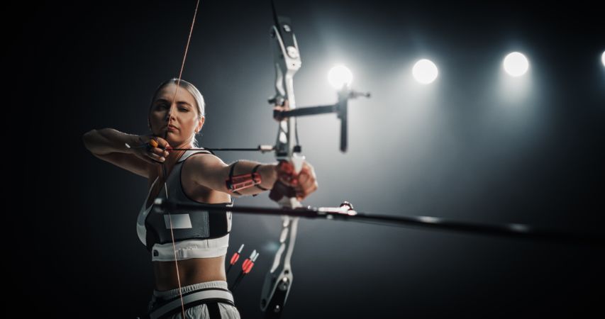 Portrait of Professional Female Archer Aiming an Arrow at a Target with a Compound Bow. Strong Athlete is Fully Equipped and Protected with Chest and Arm Guards, Preparing For Tournament