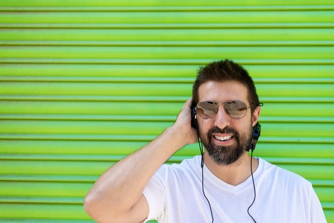 Smiling man in sunglasses listening to headphones in front of green background