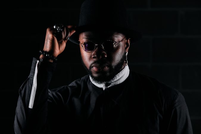 Stylish Black man touching hat and looking at camera in dark roo