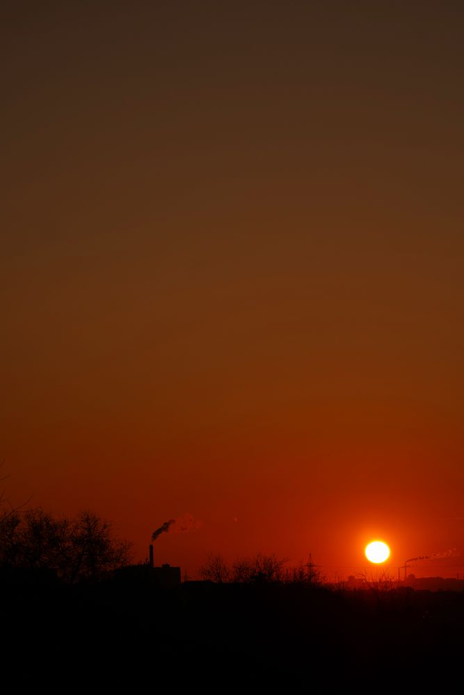 Fiery orange sun sinking behind silhouetted factories releasing smoke