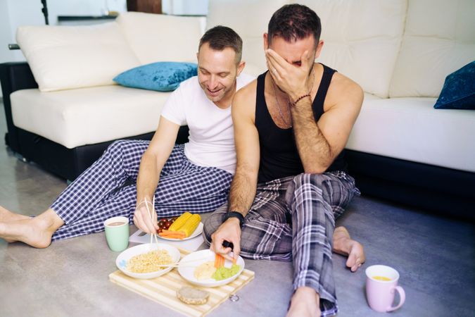 Male couple laughing while enjoying a relaxing meal at home sitting on floor