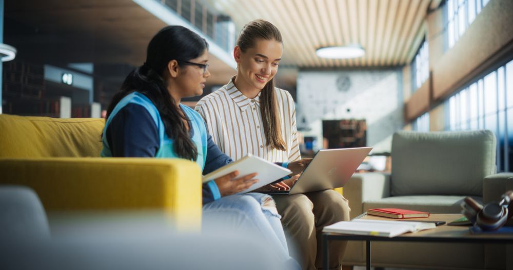 Two Diverse Friends and Classmates Working on a Collaborative Team Project in an Academic Library. Young Indian and White Women Using Laptop Computer for Online Learning and Virtual Lectures
