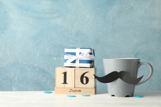 Cup with decorative mustache, gift box and wooden calendar on table against blue background, space for text