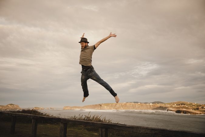Young man in hats having fun on seat on coast near waving sea and cloudy sky
