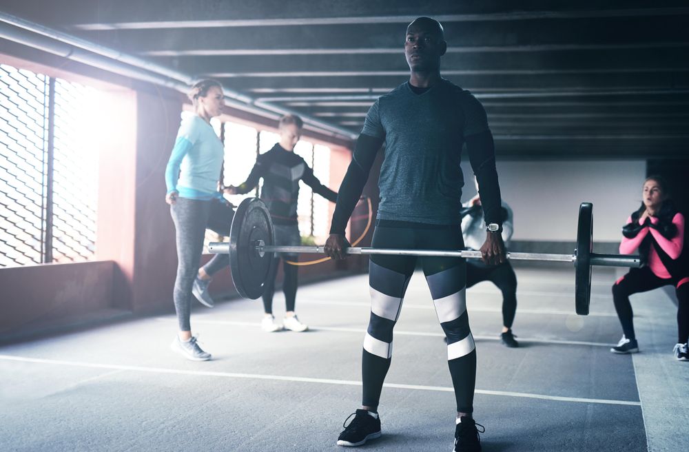 Black male with heavy barbell with people doing circuit training behind him
