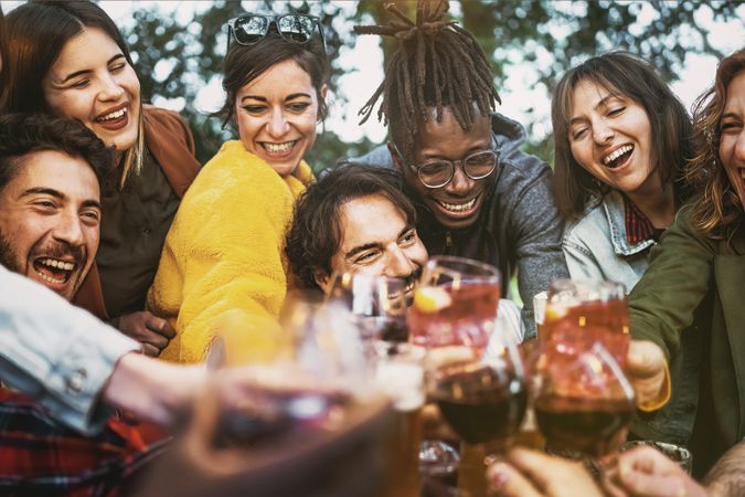 Multi-ethnic group of excited people having fun toasting wine and beers together at terrace birthday party
