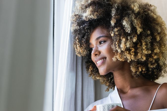 Dreamy Black woman with cup of drink near window