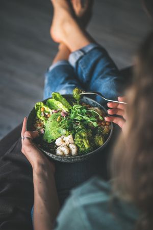 Woman in t-shirt and jeans sitting with fork in vegetarian bowl, vertical composition, top view