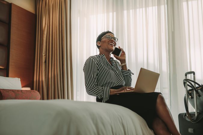 Happy businesswoman talking on mobile phone while sitting on the hotel room bed with laptop