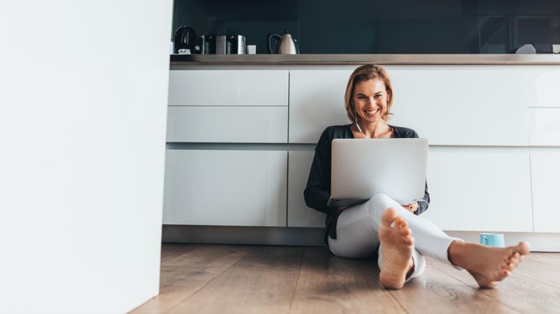 Woman with laptop sitting on the kitchen floor wearing earphones