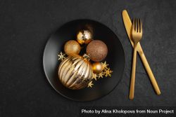 Gold Christmas decorations in dark bowl on table with gold cutlery 0JwAn5
