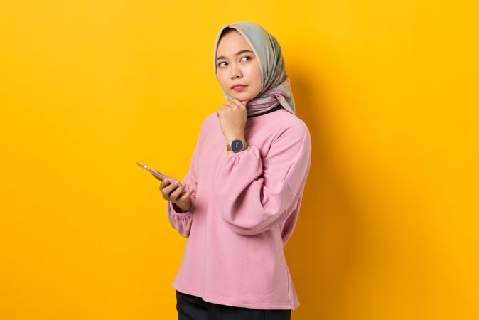 Muslim woman in headscarf holding smart phone and looking away with hand on her chin