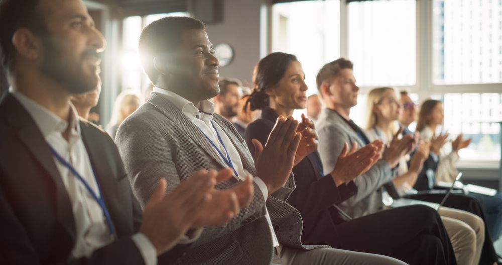 Black Man Sitting in Crowded Auditorium at a Tech Conference. Black Businessman Listening To Keynote. Specialist Watching Innovative Technology Presentation And Clapping With Audience.