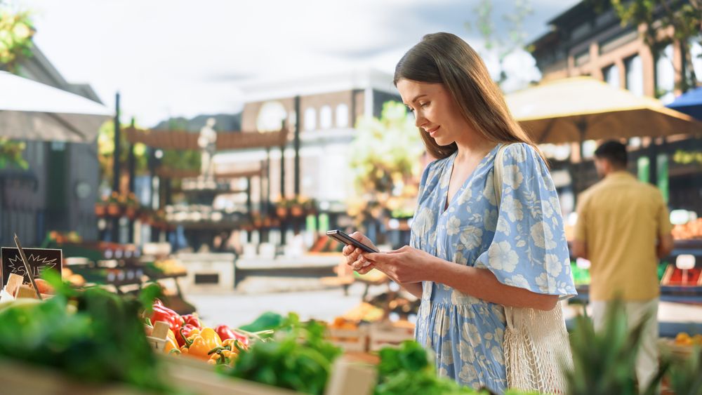 Customer in a Light Blue Dress Shopping for Fresh Seasonal Fruits and Vegetables, Using Smartphone