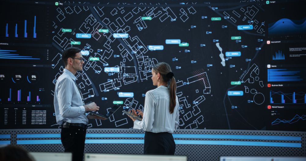 Male And Female Data Analysts Discussing New Locations For Business In Front Of Big Digital Screen With City Map In Monitoring Office.