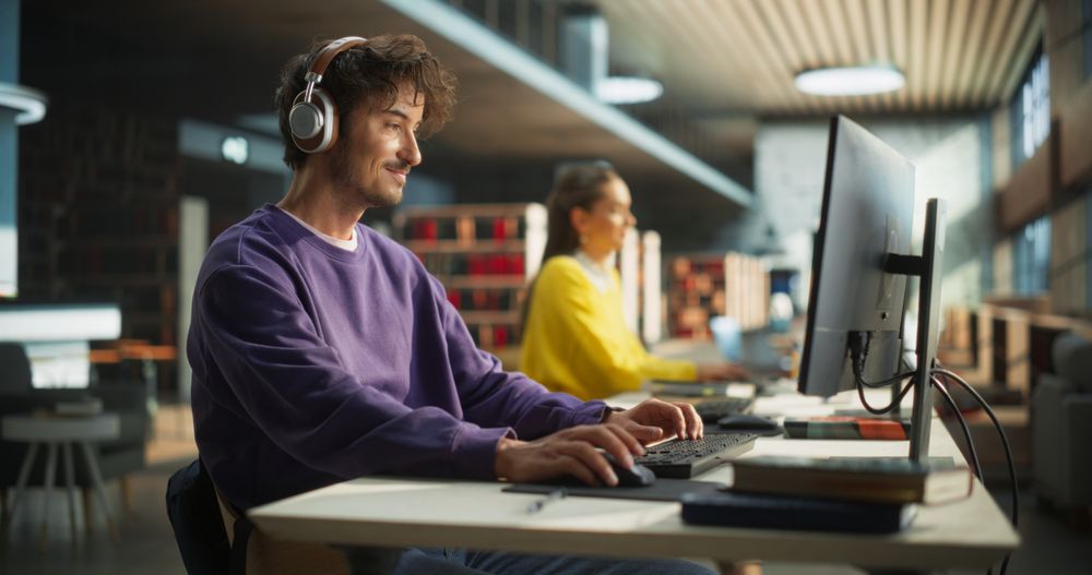 Excited Young Male is Focused on Creating a College Presentation for Class Assignment, Using Online Office Software on Desktop Computer in a Public Library Space with Other Students
