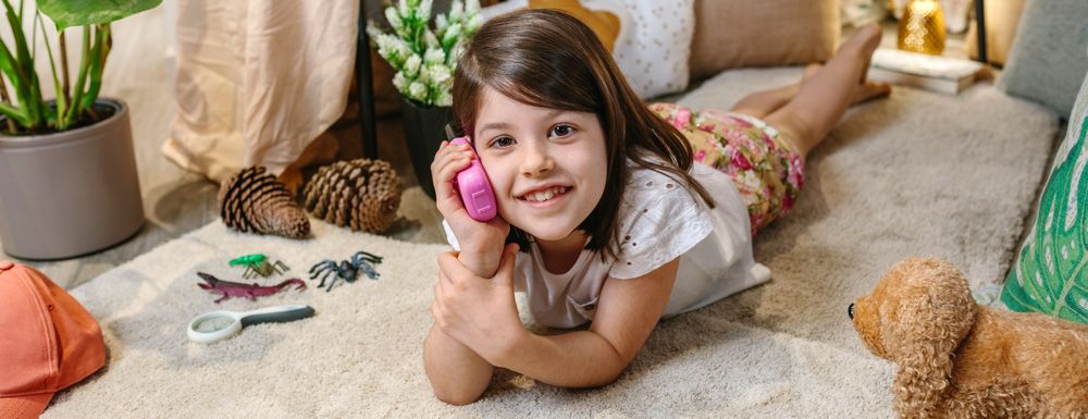 Banner of smiling little girl looking at camera while using walkie-talkie lying over rug at home