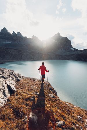 Back view of man in red jacket with hiking sticks standing by lake