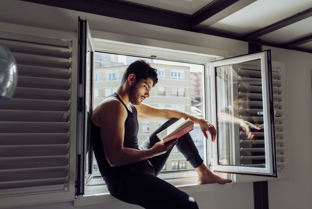 Young male sitting on window and reading book
