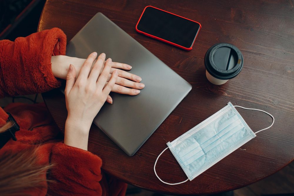 Top view of woman in red jacket sitting at a table with laptop and phone and cup of coffee along with facemask
