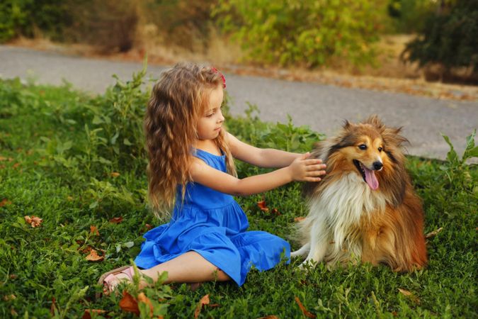 Adorable child in blue dress petting dog in the grass