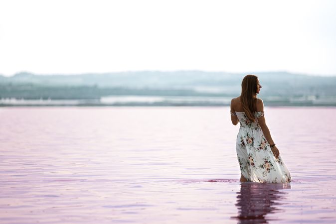 Peaceful woman in white dress walking ankle deep in pink lake