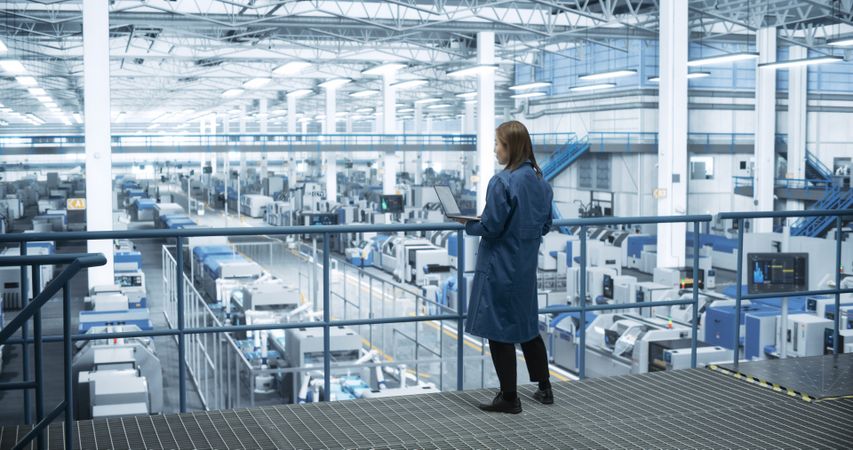Young Asian Female Engineer Standing on a Platform, Using Laptop Computer at an Electronics Factory. Machines are Undergoing Maintenance, Specialist Monitoring the Progress Through Online Software