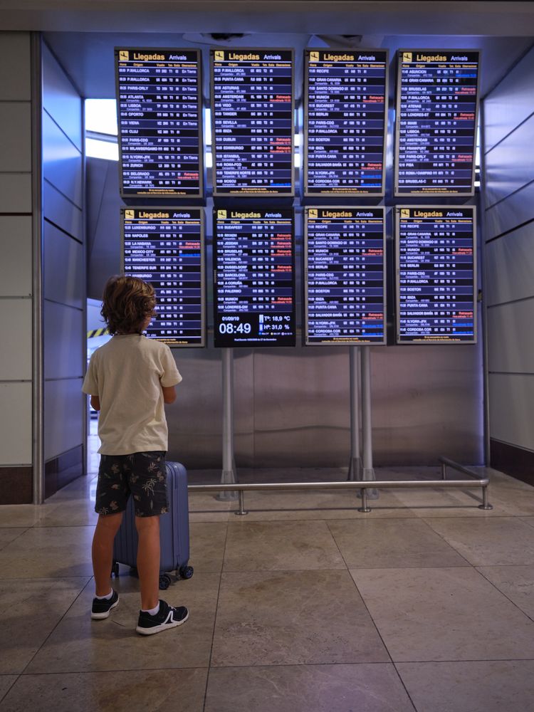 Unrecognizable boy with blue suitcase looking at arrival boards