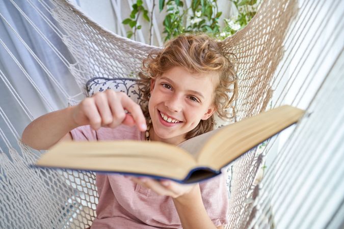 Positive boy lying in hammock and pointing at book
